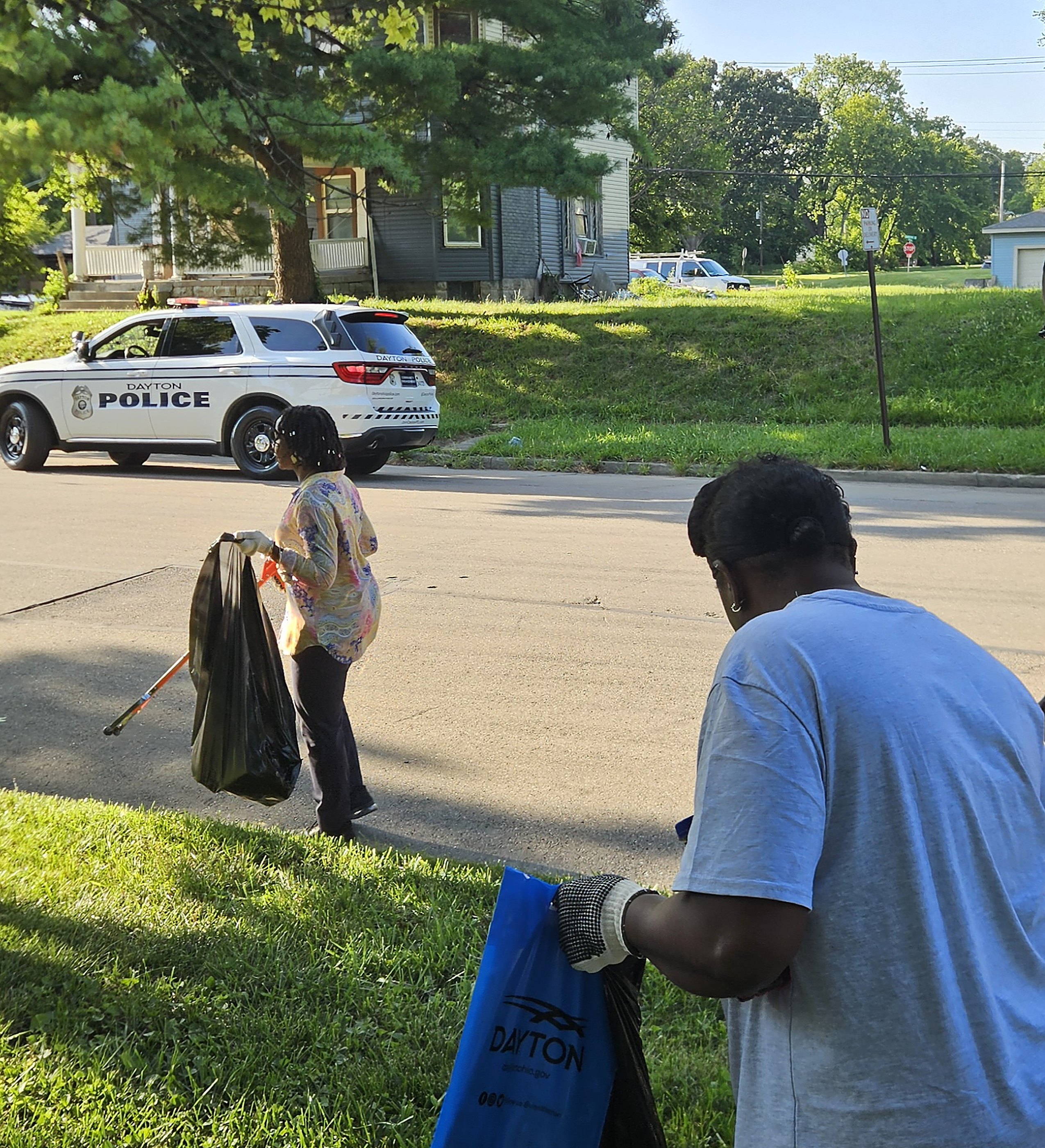 A Dayton volunteer with a litter grabber cleaning a residential street, a police cruiser and neighbors in the background